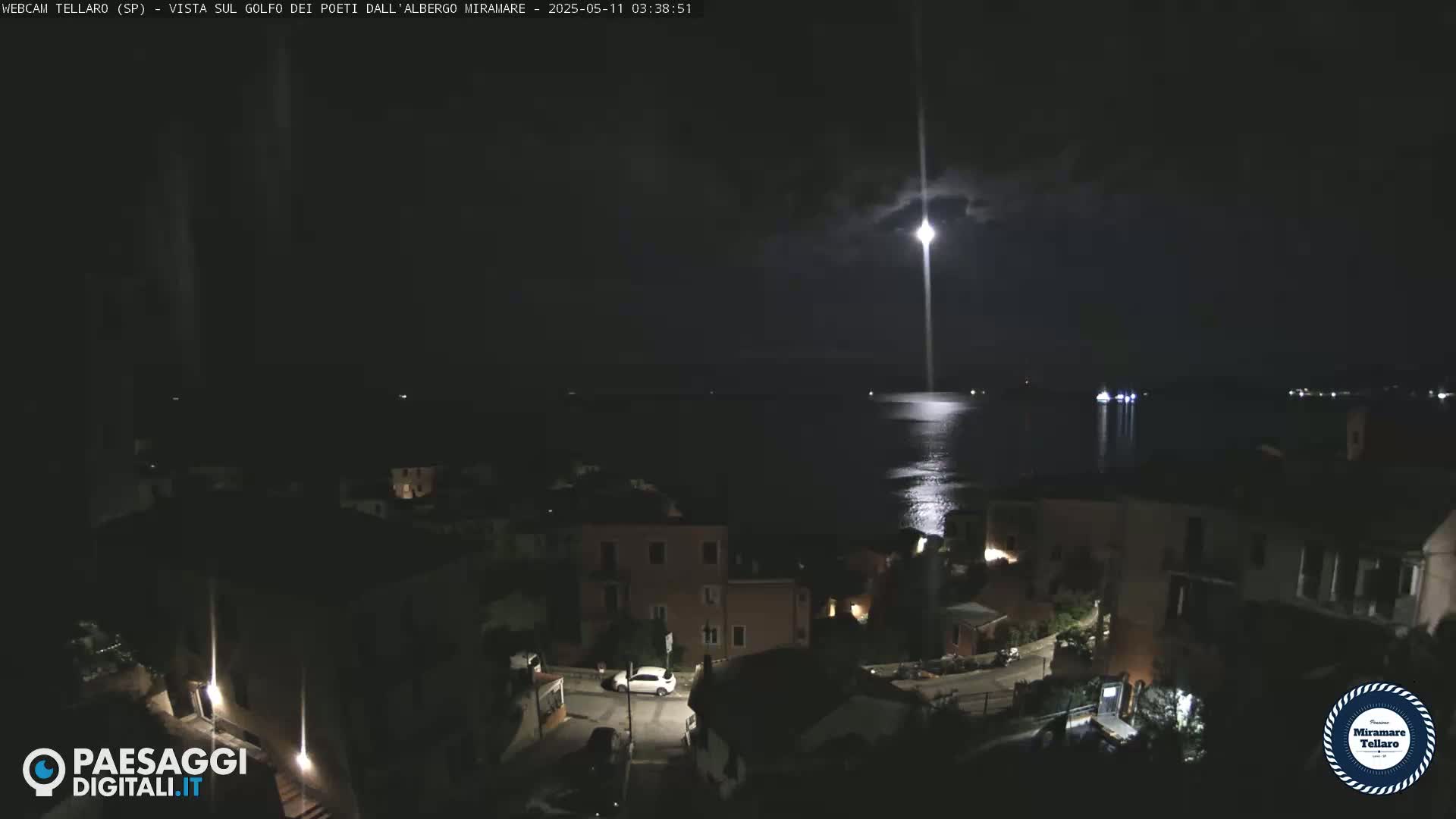 A nighttime coastal town is shown, illuminated by streetlights and a bright moon, overlooking a dark body of water.
