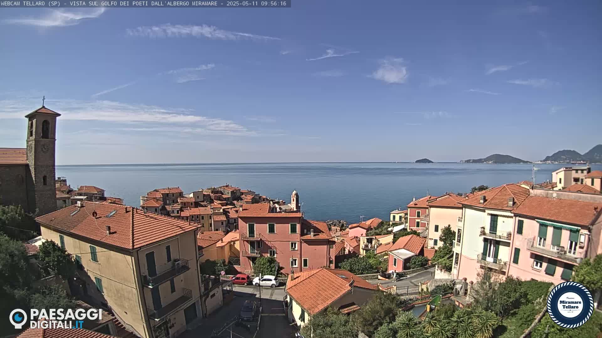 A coastal town with red-tiled roofs and a bell tower overlooks a calm sea under a mostly clear blue sky.