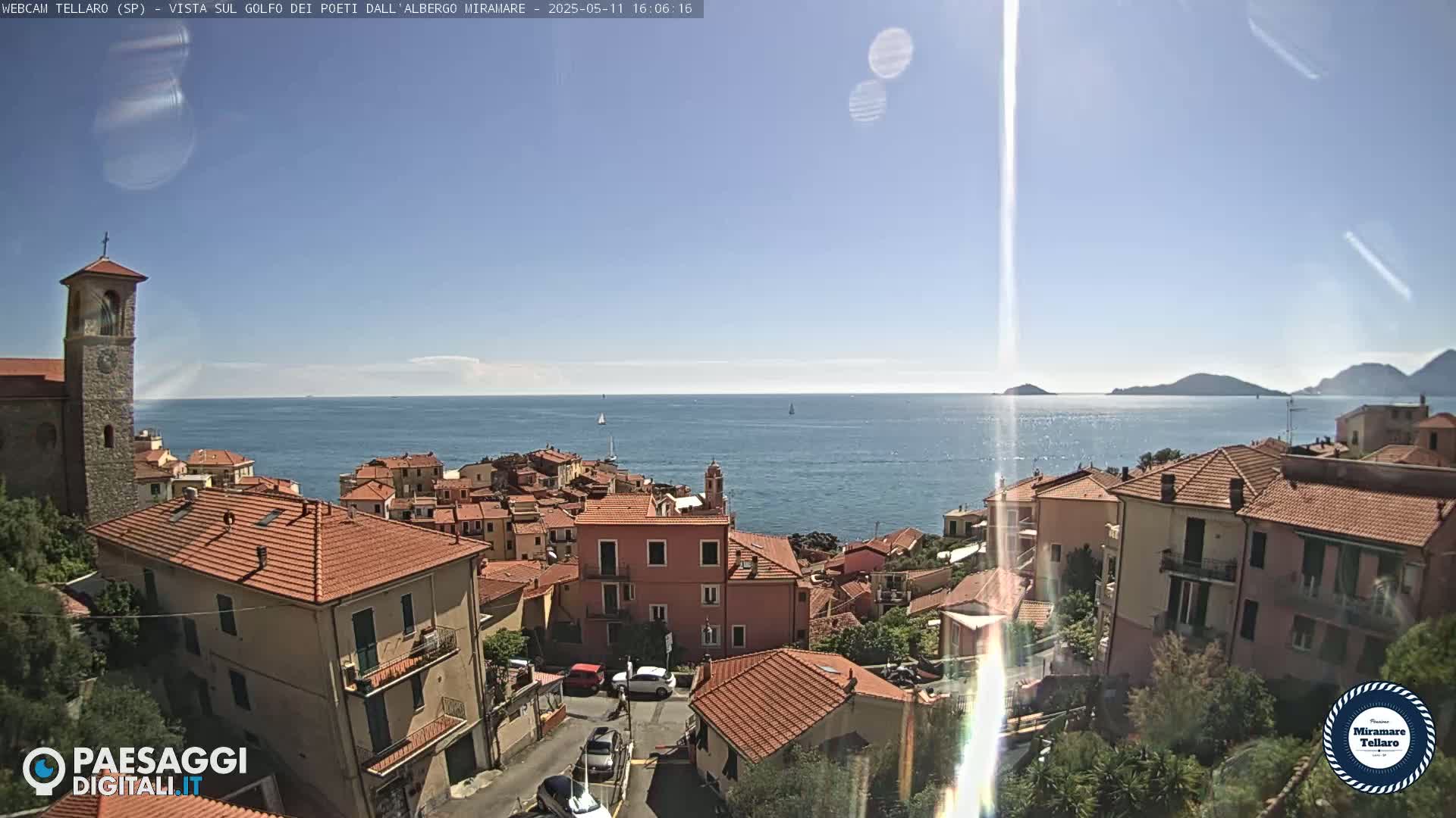A coastal town with red-tiled roofs and a stone church overlooks a calm sea under a sunny sky.