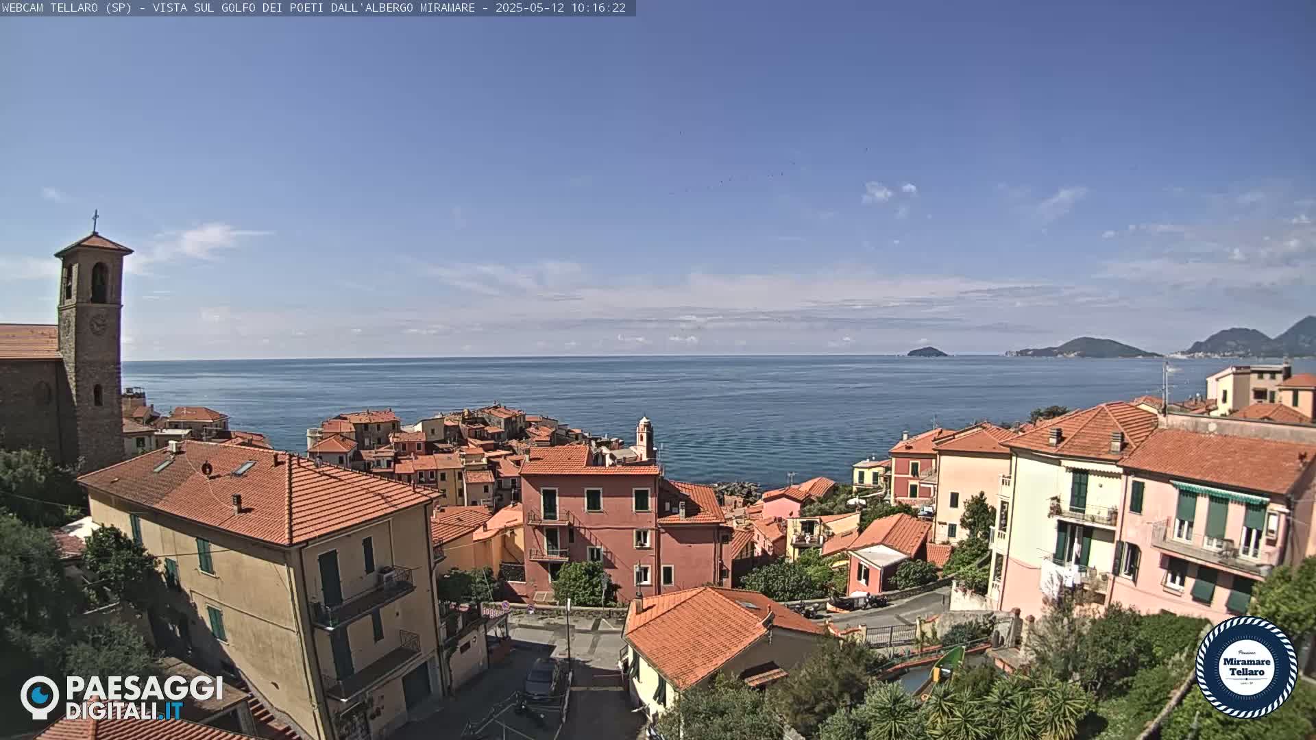 A coastal town with red-tiled roofs overlooks a calm blue sea under a mostly clear sky.