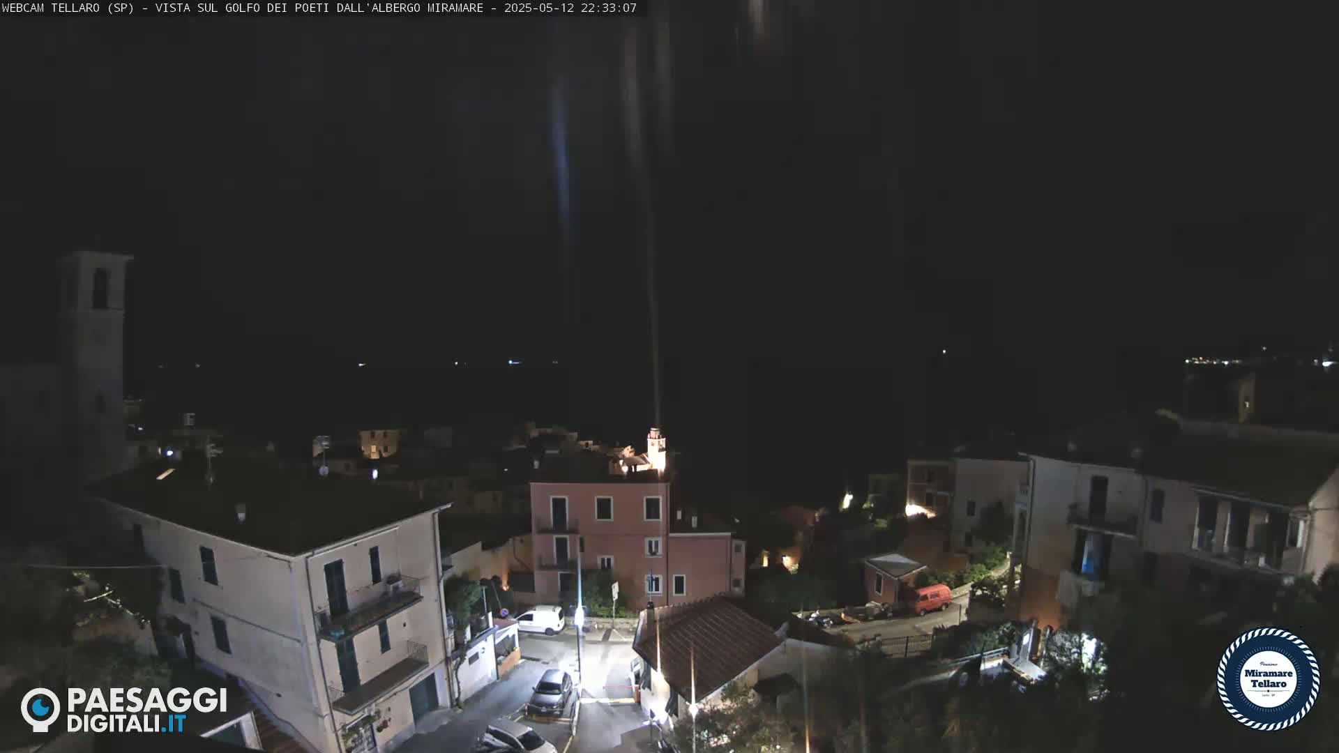 A nighttime, high-angle view of a small town nestled on a hillside, with a beam of light extending upward into the dark sky.