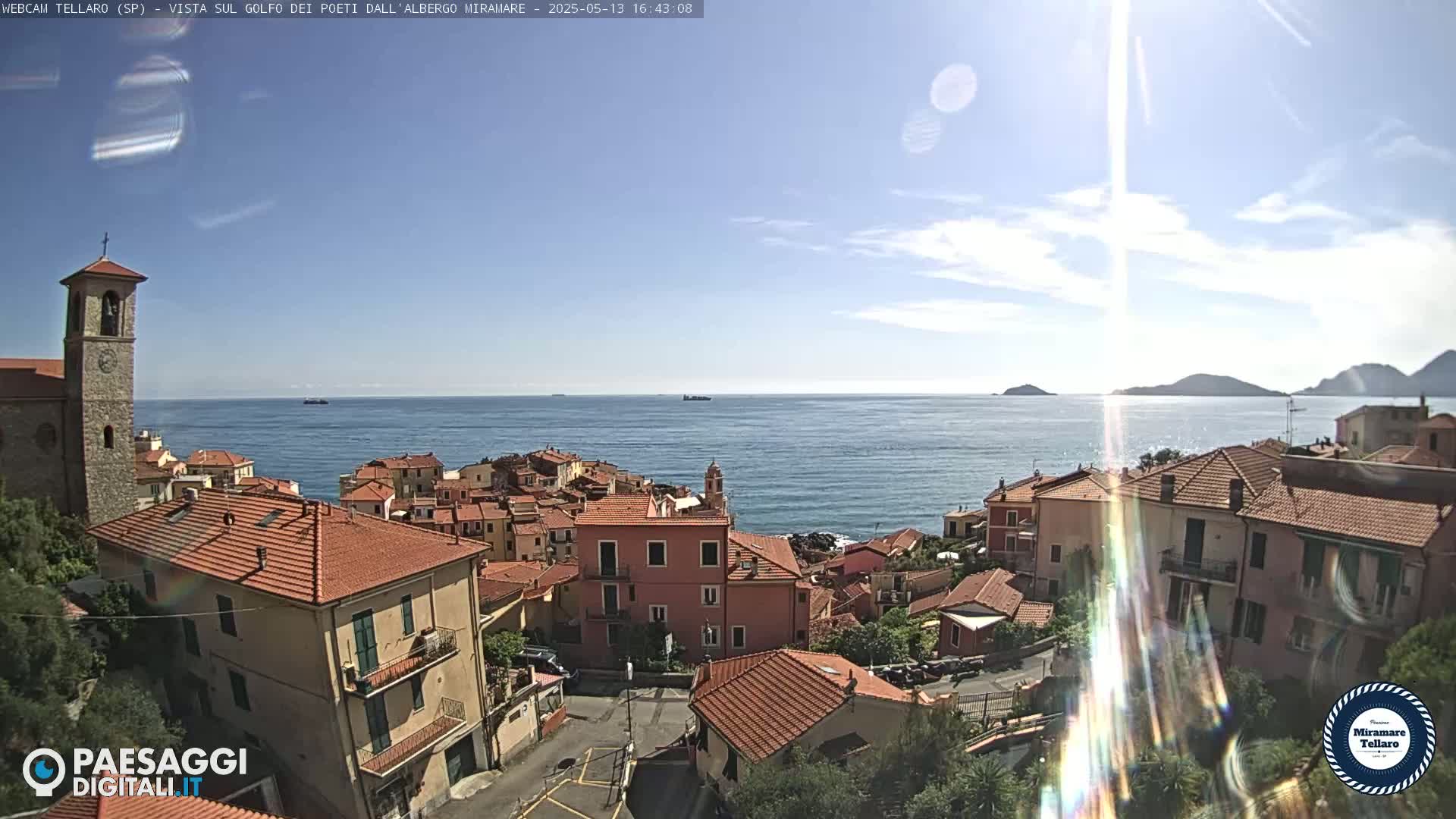 A coastal town with red-tiled roofs overlooks a calm sea under a mostly sunny sky.