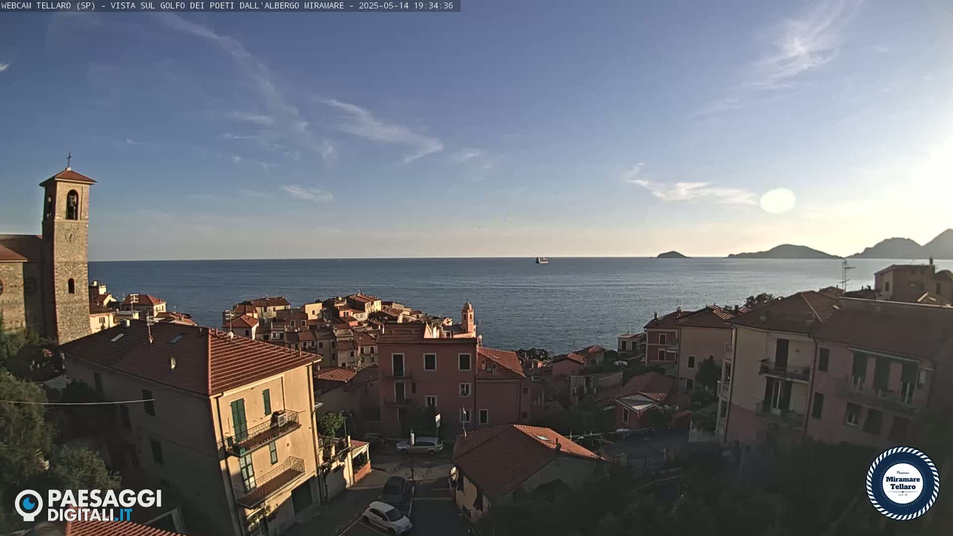 A coastal town with red-tiled roofs and a church steeple overlooks a calm sea under a mostly clear, sunny sky.