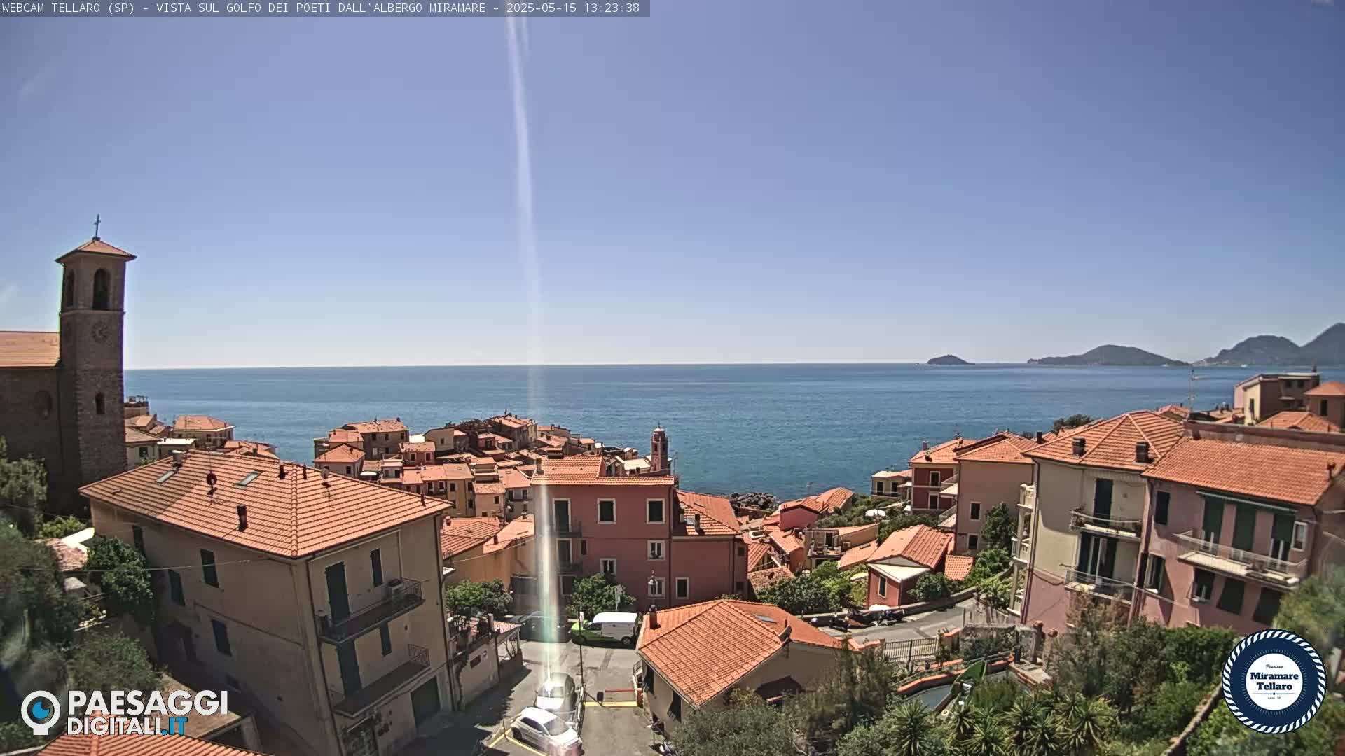 A coastal town with red-tiled roofs and a church steeple overlooks a calm blue sea under a clear, sunny sky.