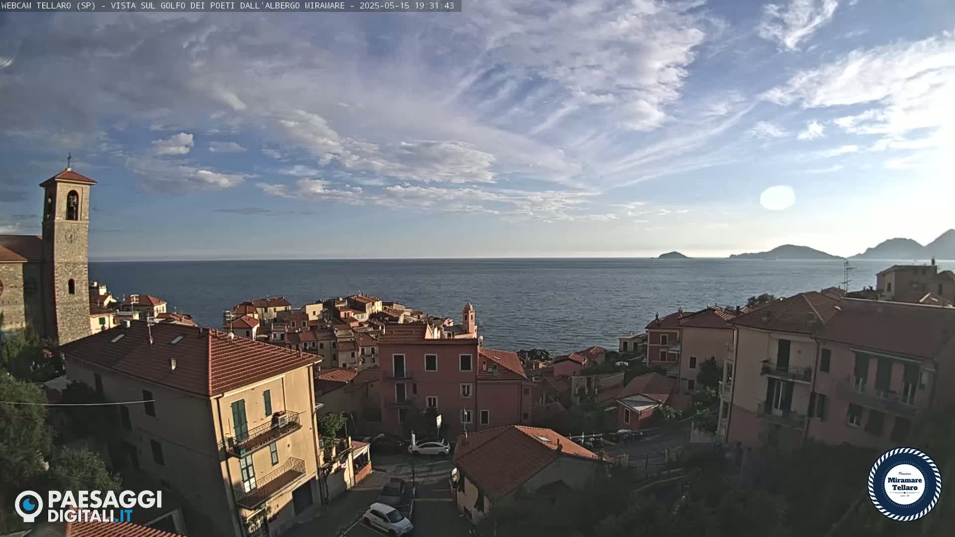 A coastal town with red-tiled rooftops, a church steeple, and a calm ocean under a partly cloudy sky.