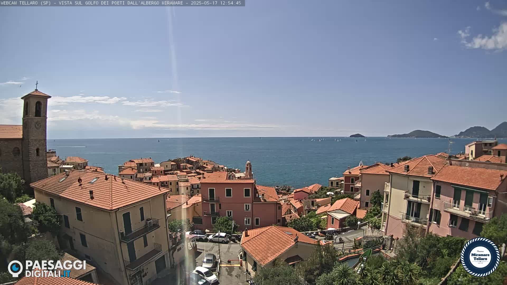 A coastal town with red-tiled roofs and a bell tower overlooks a calm blue sea dotted with sailboats under a mostly clear sky.