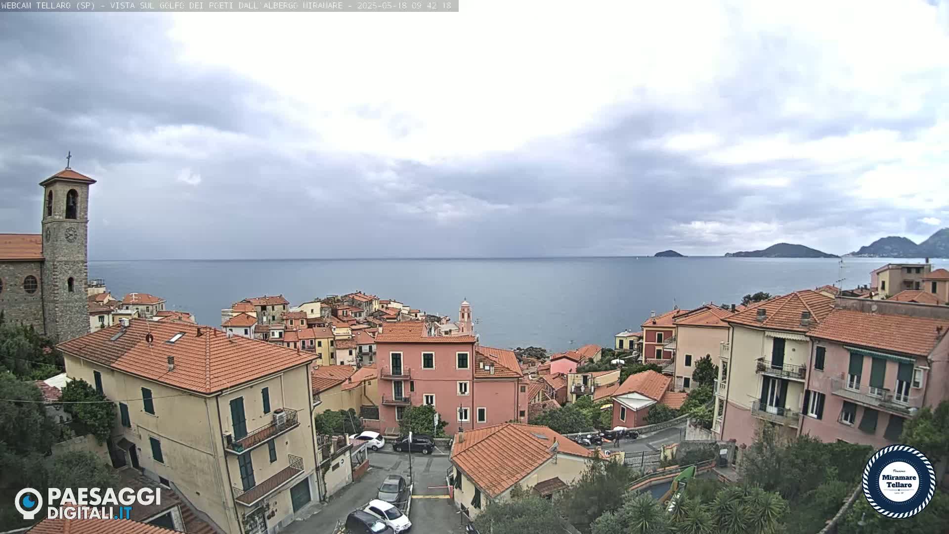 A coastal town with red-tiled roofs overlooks a calm sea under a cloudy sky.