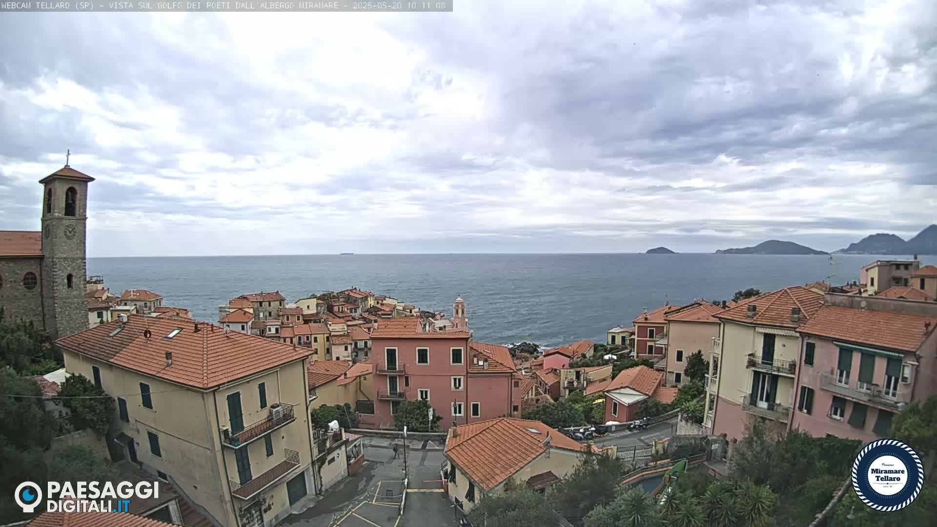 A coastal town with red-tiled roofs and a church steeple overlooks a calm sea under a mostly cloudy sky.