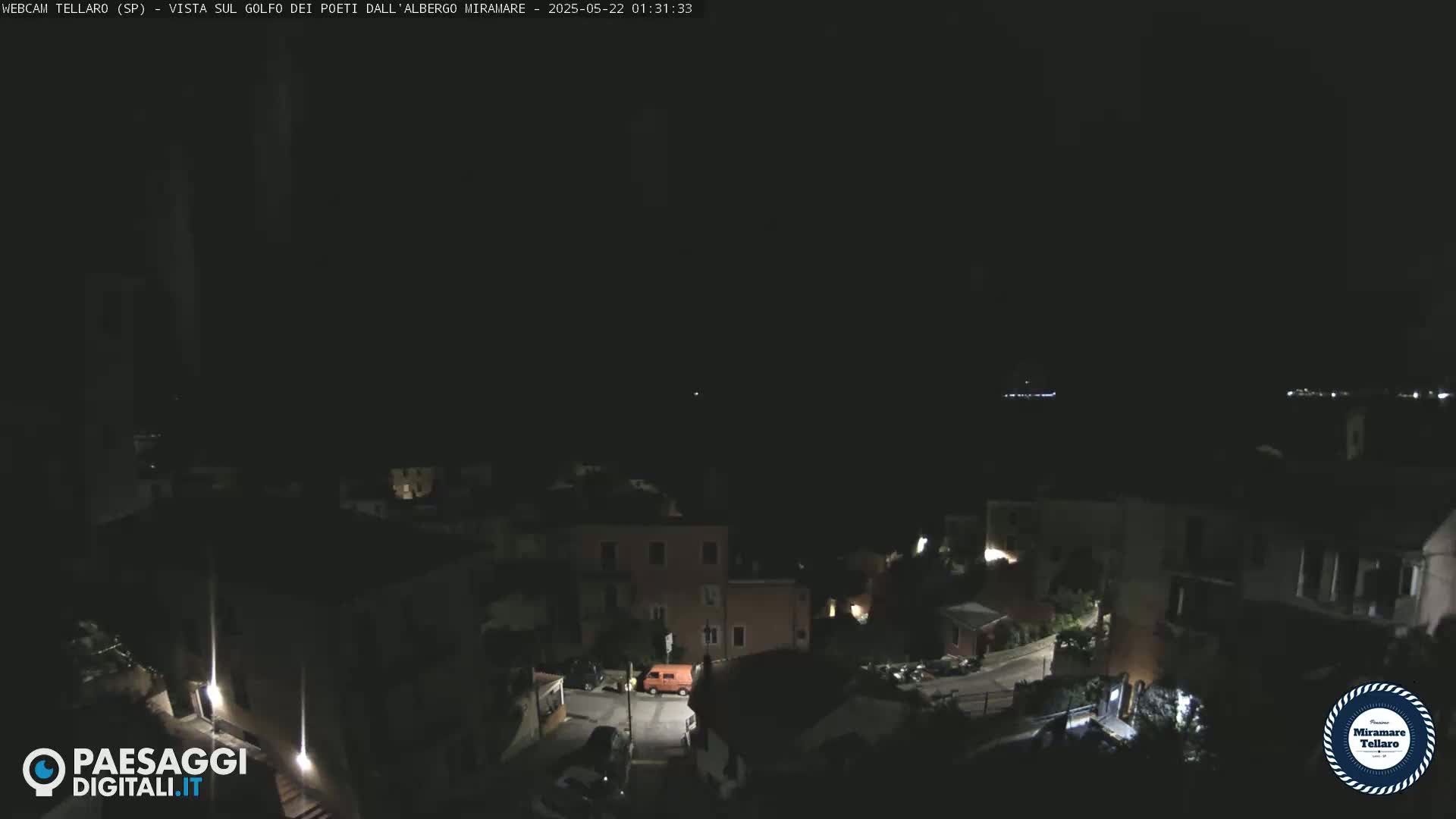 A nighttime view of a small town nestled on a hillside, with dimly lit buildings and a few cars visible, under a dark sky.