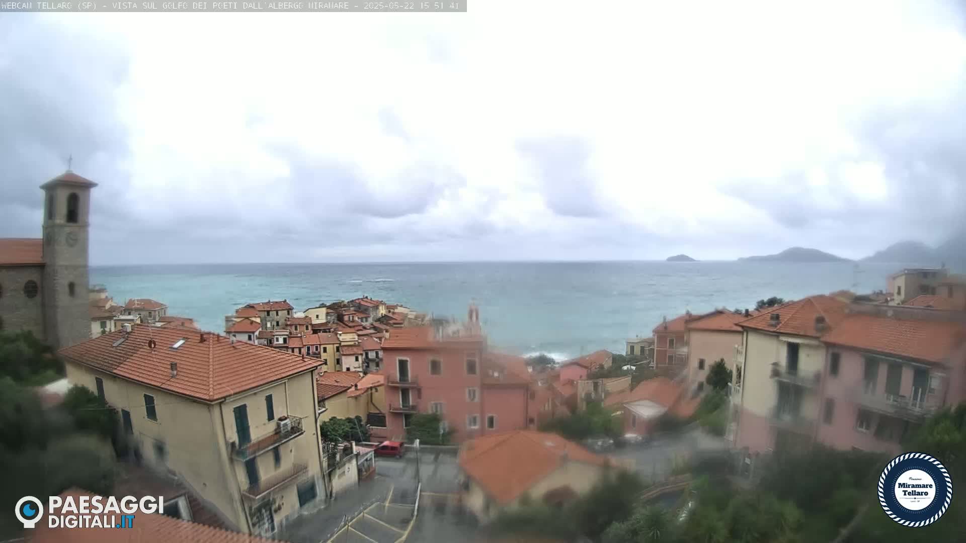 A seaside town with red-tiled roofs and a church steeple is visible under an overcast sky, with a choppy sea in the background.