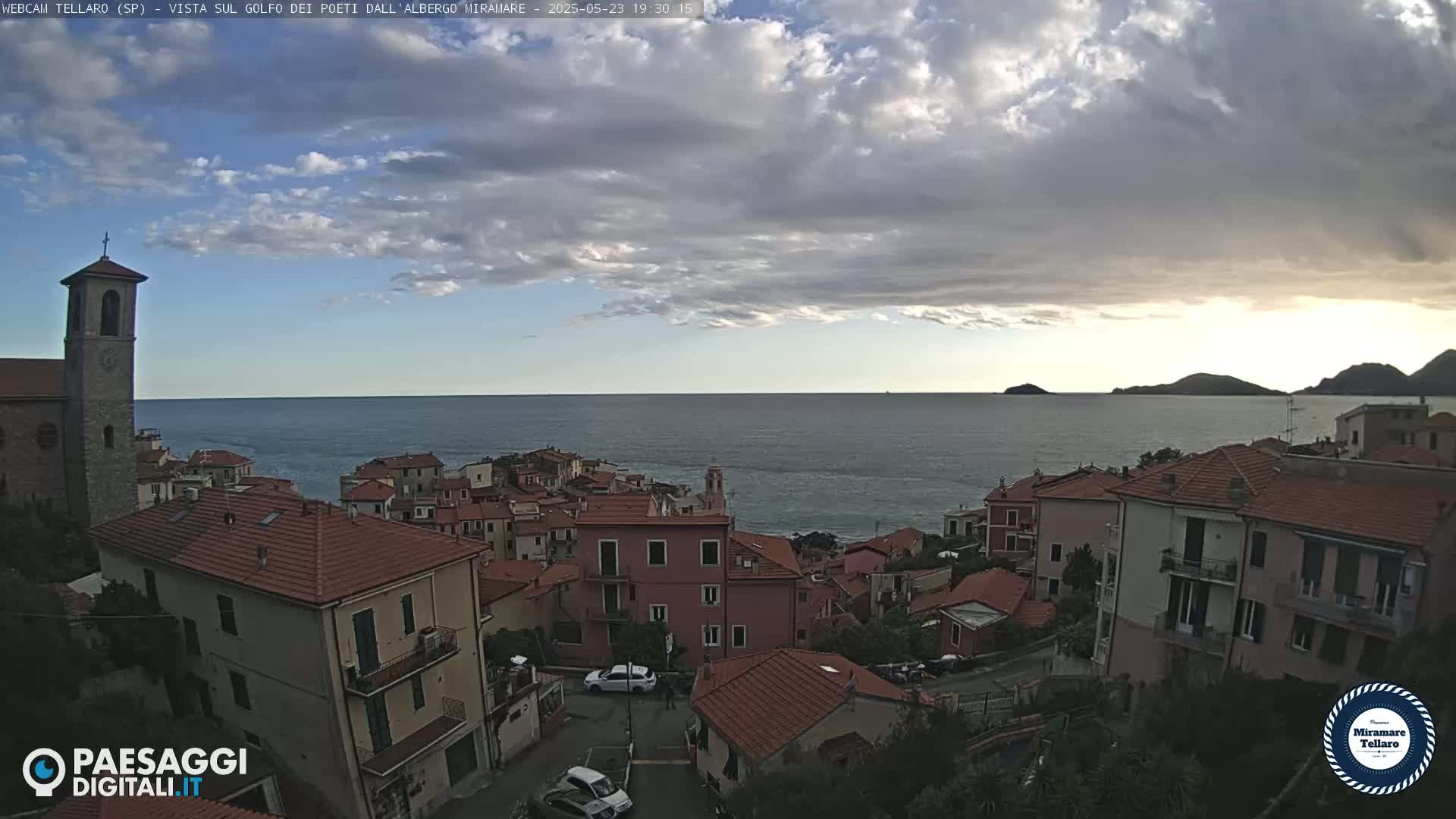 A coastal town with red-tiled roofs overlooks a calm sea under a partly cloudy sky.