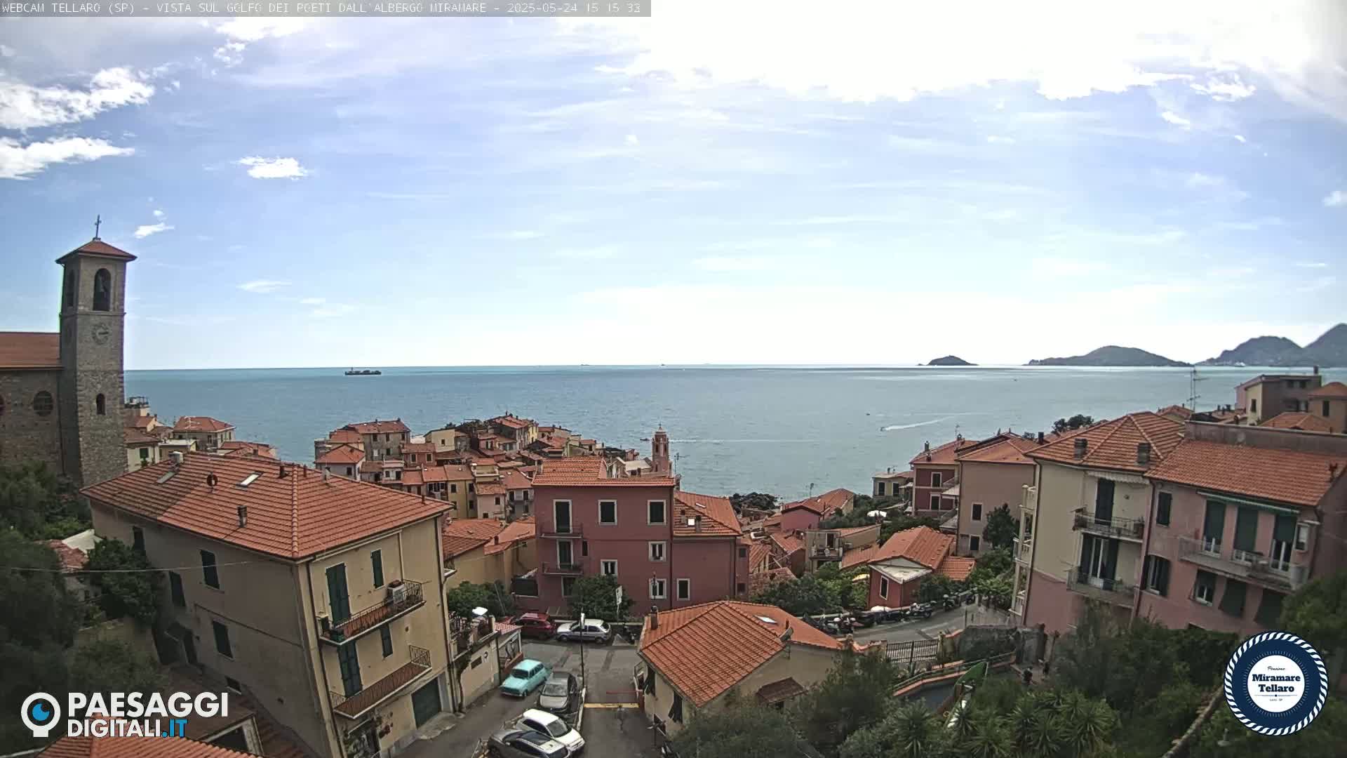 A coastal town with red-tiled roofs and a church tower overlooks a calm sea under a partly cloudy sky.