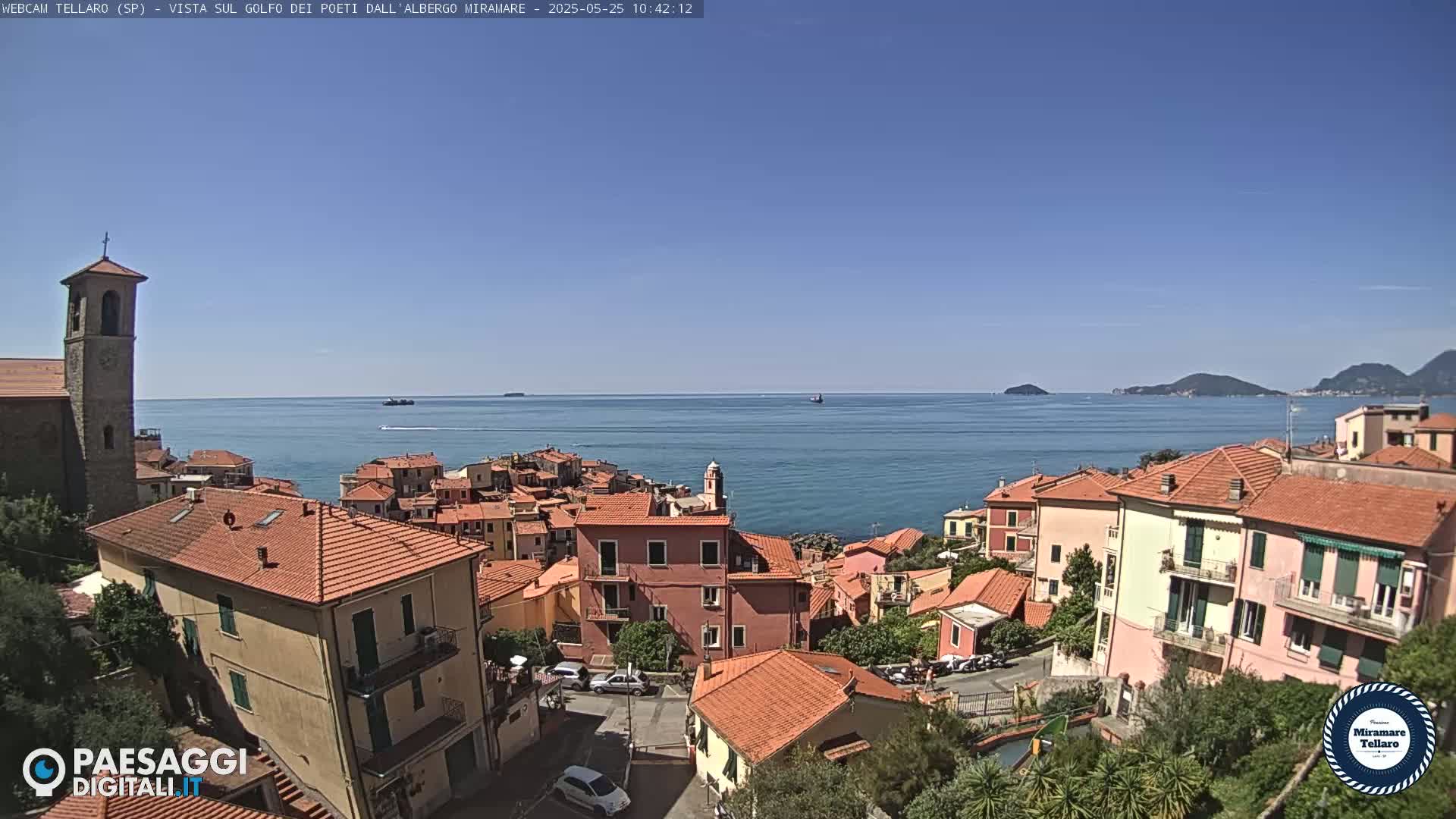 A coastal town with red-tiled roofs and a bell tower overlooks a calm blue sea under a clear blue sky.