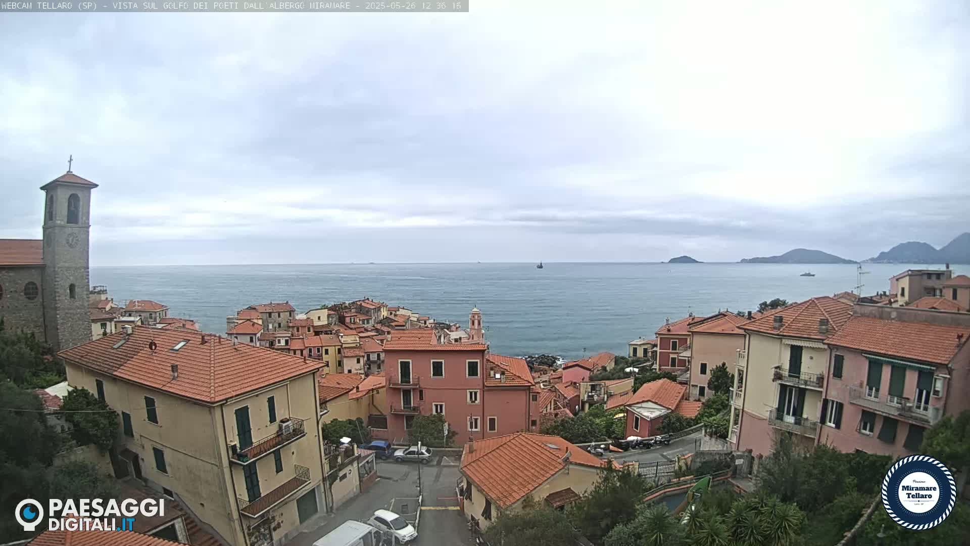 A coastal town with red-tiled roofs and a church steeple overlooks a calm sea under a cloudy sky.