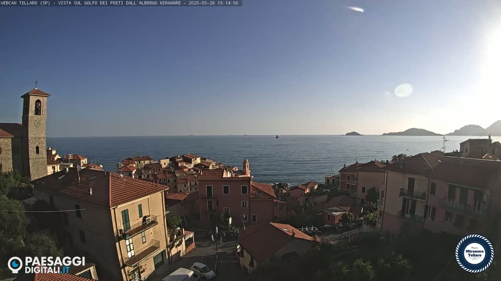A coastal town with red-tiled roofs and a church steeple overlooks a calm sea under a clear, sunny sky.