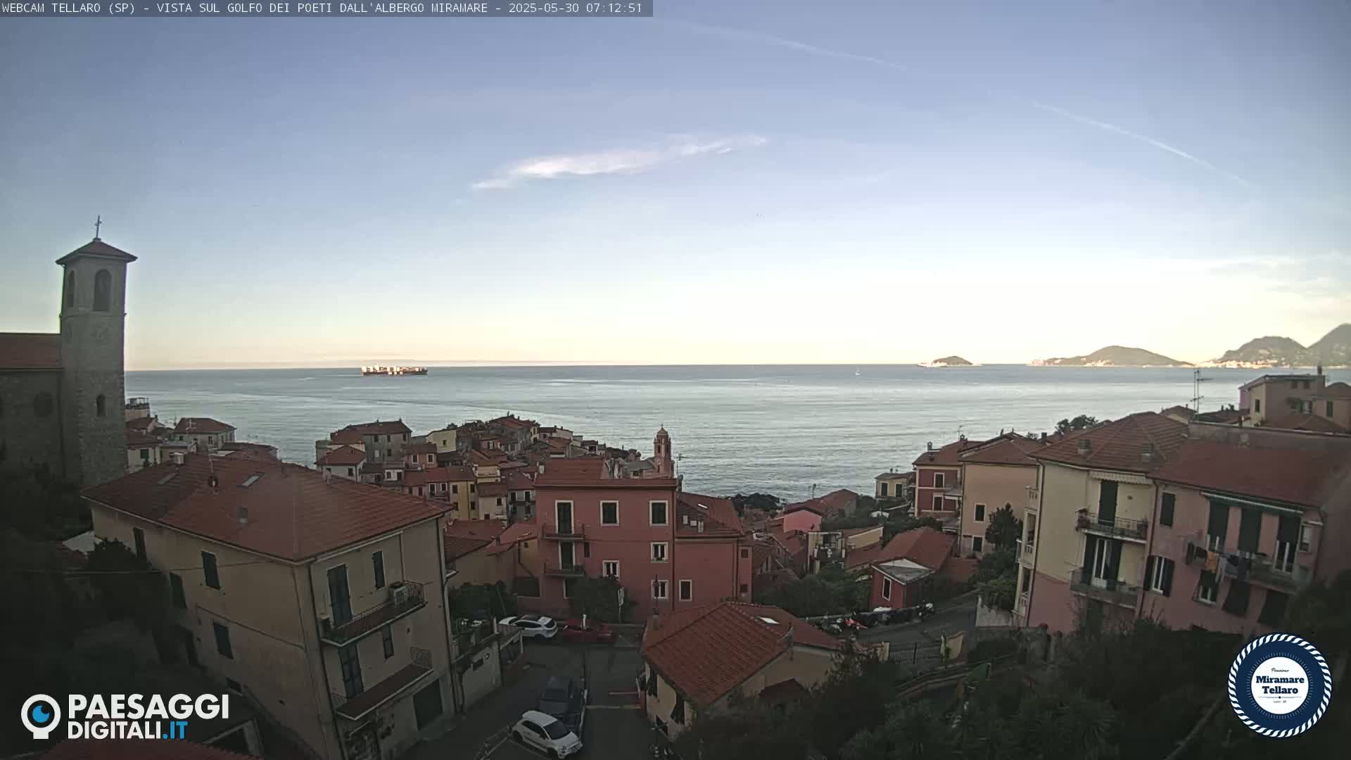 A coastal town with red-tiled roofs overlooks a calm sea under a clear, light blue sky.