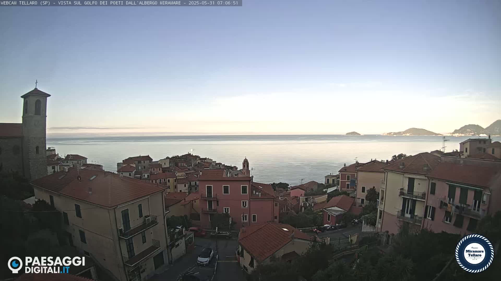 A coastal town with red-tiled roofs overlooks a calm sea under a clear, light-blue sky.