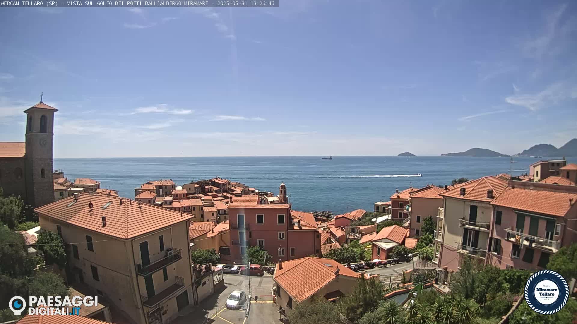 A coastal town with red-tiled rooftops and a church steeple overlooks a calm blue sea under a mostly clear sky.