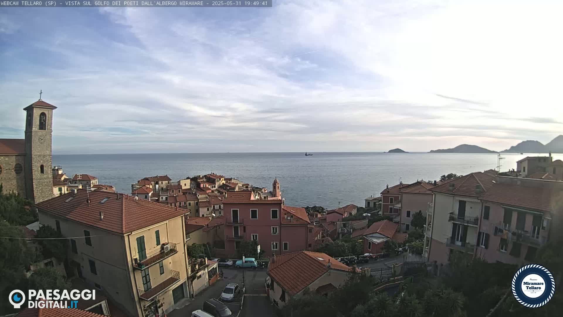 A coastal town with red-tiled roofs and a church steeple overlooks a calm sea under a partly cloudy sky.