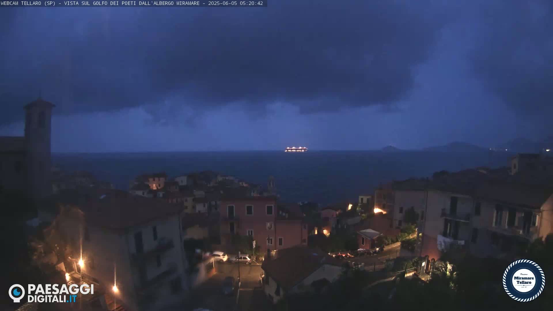 A coastal town at night under a dark, cloudy sky, with a ship visible on the dark sea in the distance.