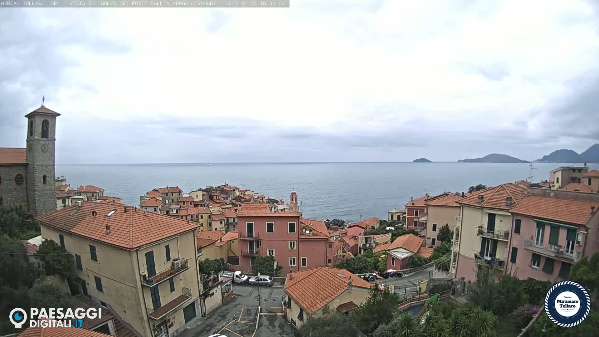 A coastal town with red-tiled roofs and a church steeple overlooks a calm sea under an overcast sky.