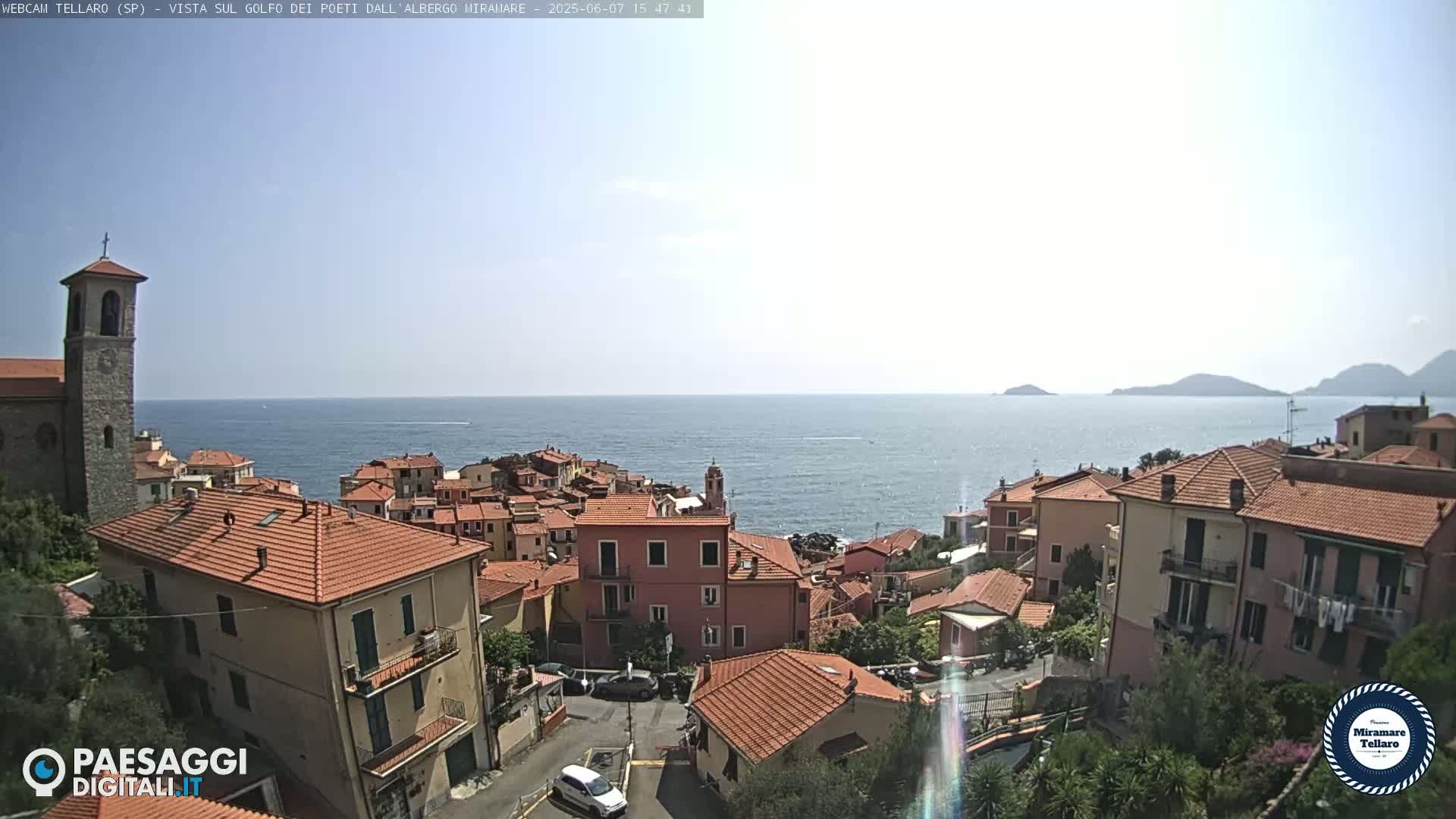 A coastal town with red-tiled roofs overlooks a calm sea under a bright, sunny sky.