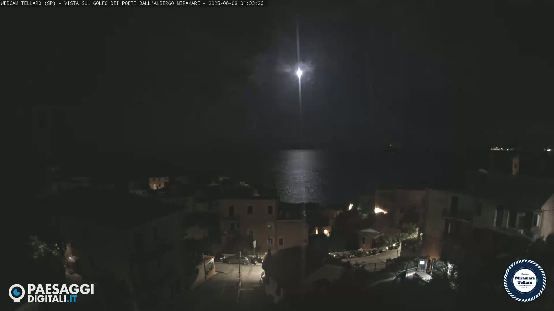 A bright light streaks across a night sky over a dark coastal town and calm sea.