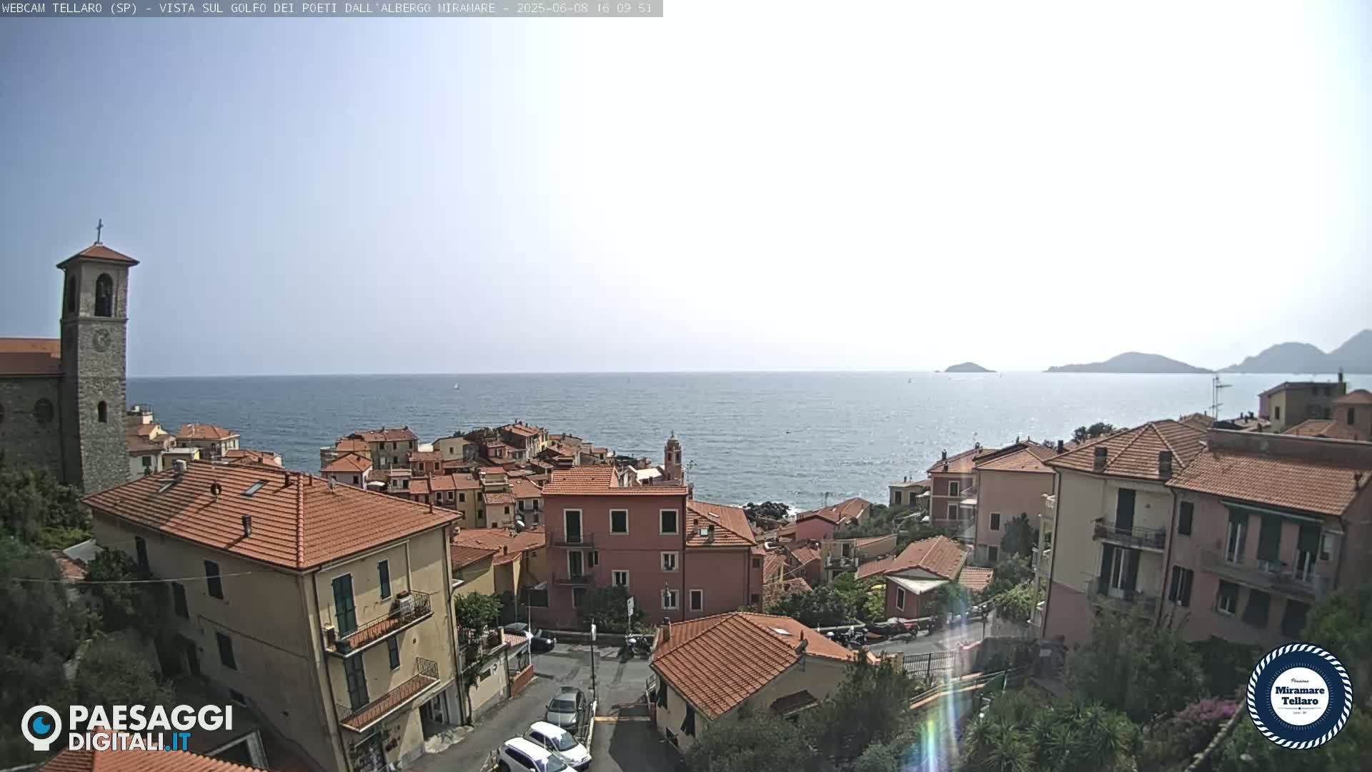 A coastal town with red-tiled roofs overlooks a calm sea under a bright, sunny sky.