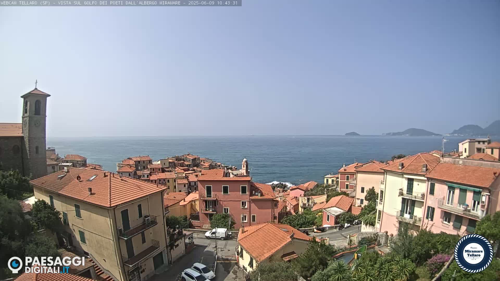 A coastal town with red-tiled roofs overlooks a calm blue sea and distant islands under a clear sky.
