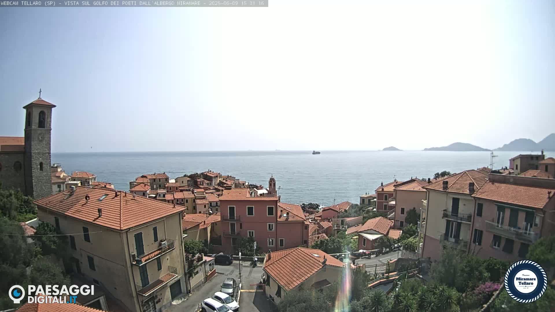 A coastal town with red-tiled roofs and a church steeple overlooks a calm sea under a bright, sunny sky.