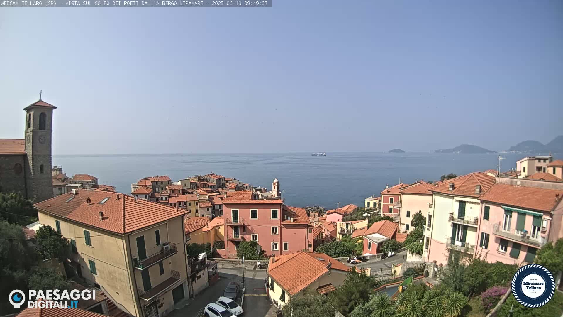 A coastal town with red-tiled roofs overlooks a calm blue sea on a clear, sunny day.
