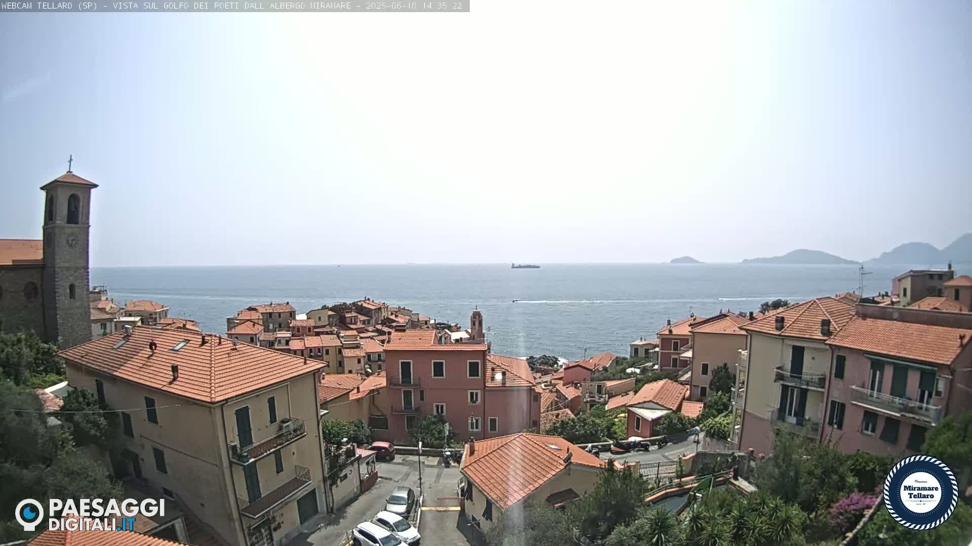 A coastal town with red-roofed buildings overlooks a calm sea under a hazy sky.
