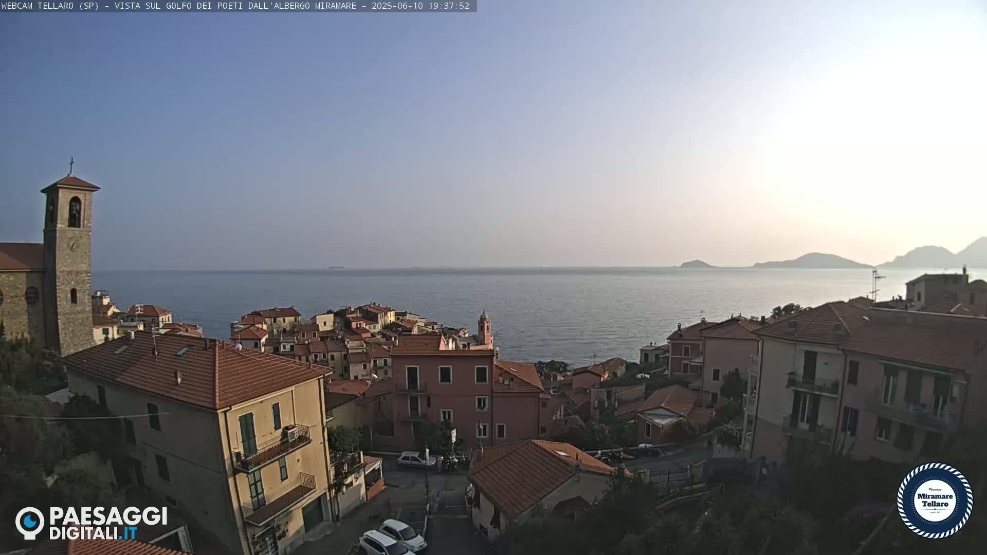 A coastal town with red-tiled roofs and a church tower overlooks a calm sea under a clear, twilight sky.