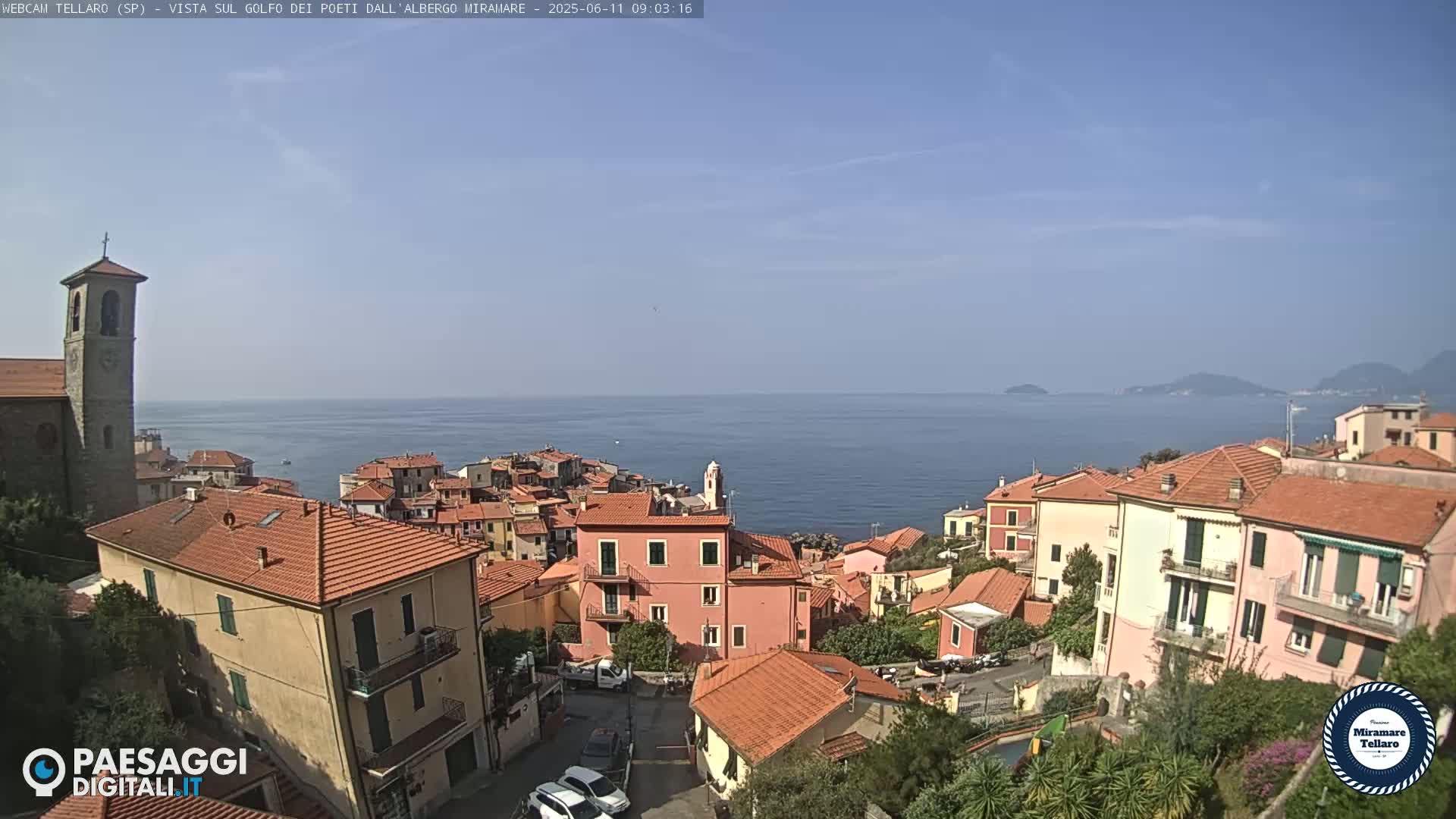 A coastal town with red-tiled roofs overlooks a calm blue sea under a clear blue sky.