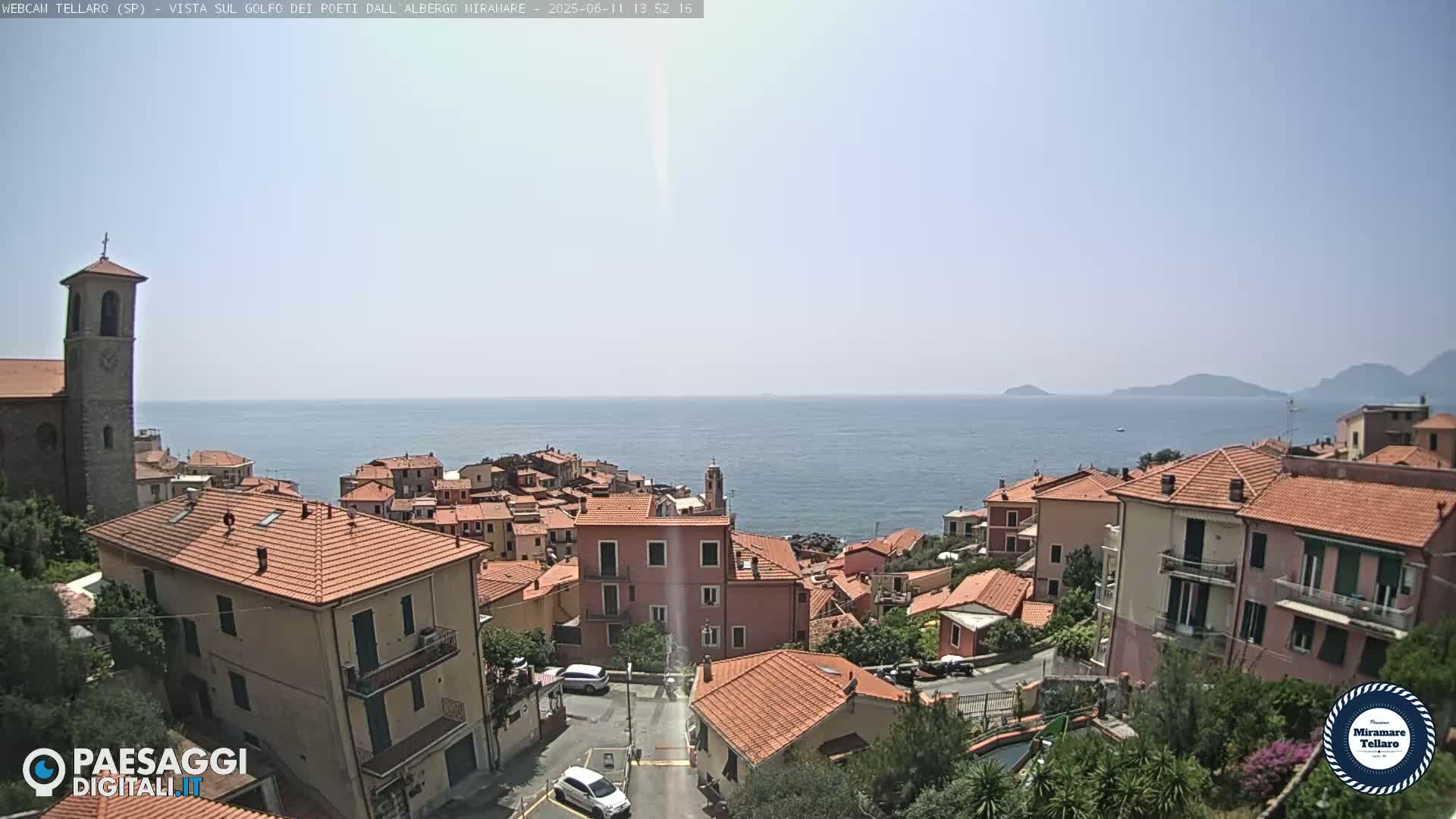 A coastal town with red-tiled roofs, a church steeple, and a clear view of the ocean under a bright, sunny sky.