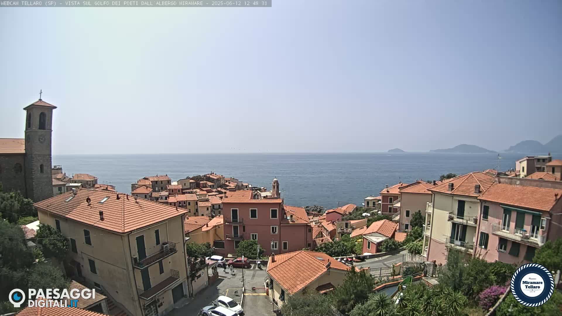 A coastal town with red-tiled roofs and a church steeple overlooks a calm blue sea under a clear sky.