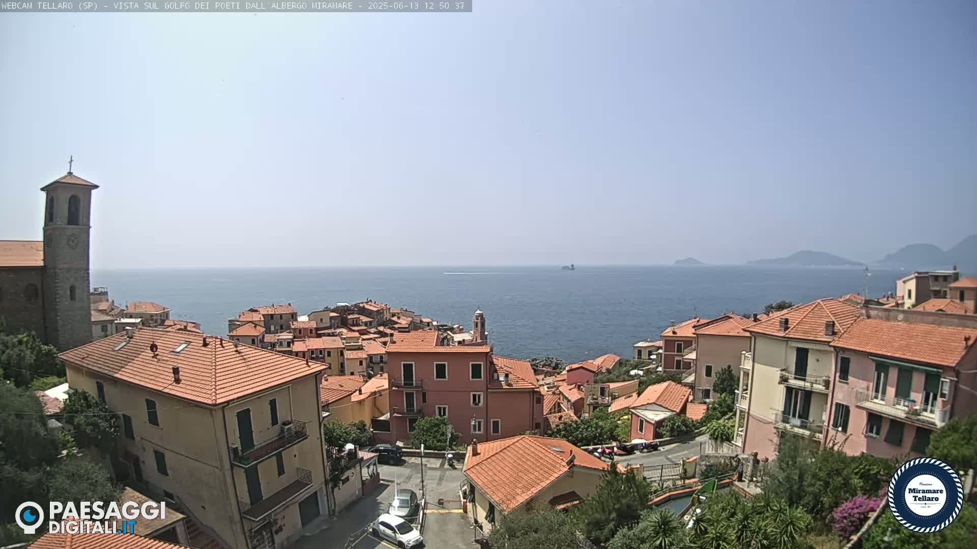 A coastal town with red-tiled roofs and a church steeple overlooks a calm, clear blue sea under a sunny sky.