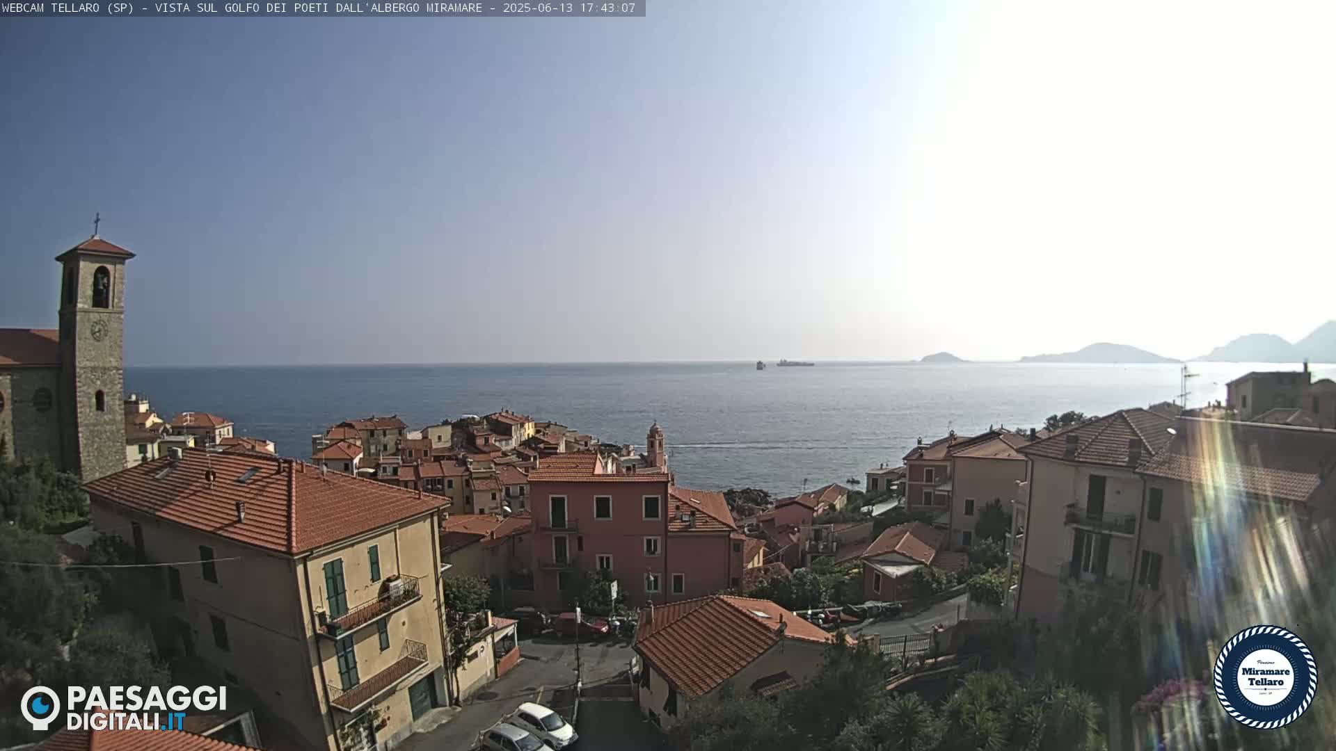 A coastal town with red-tiled roofs and a church steeple overlooks a calm, blue sea under a clear sky.