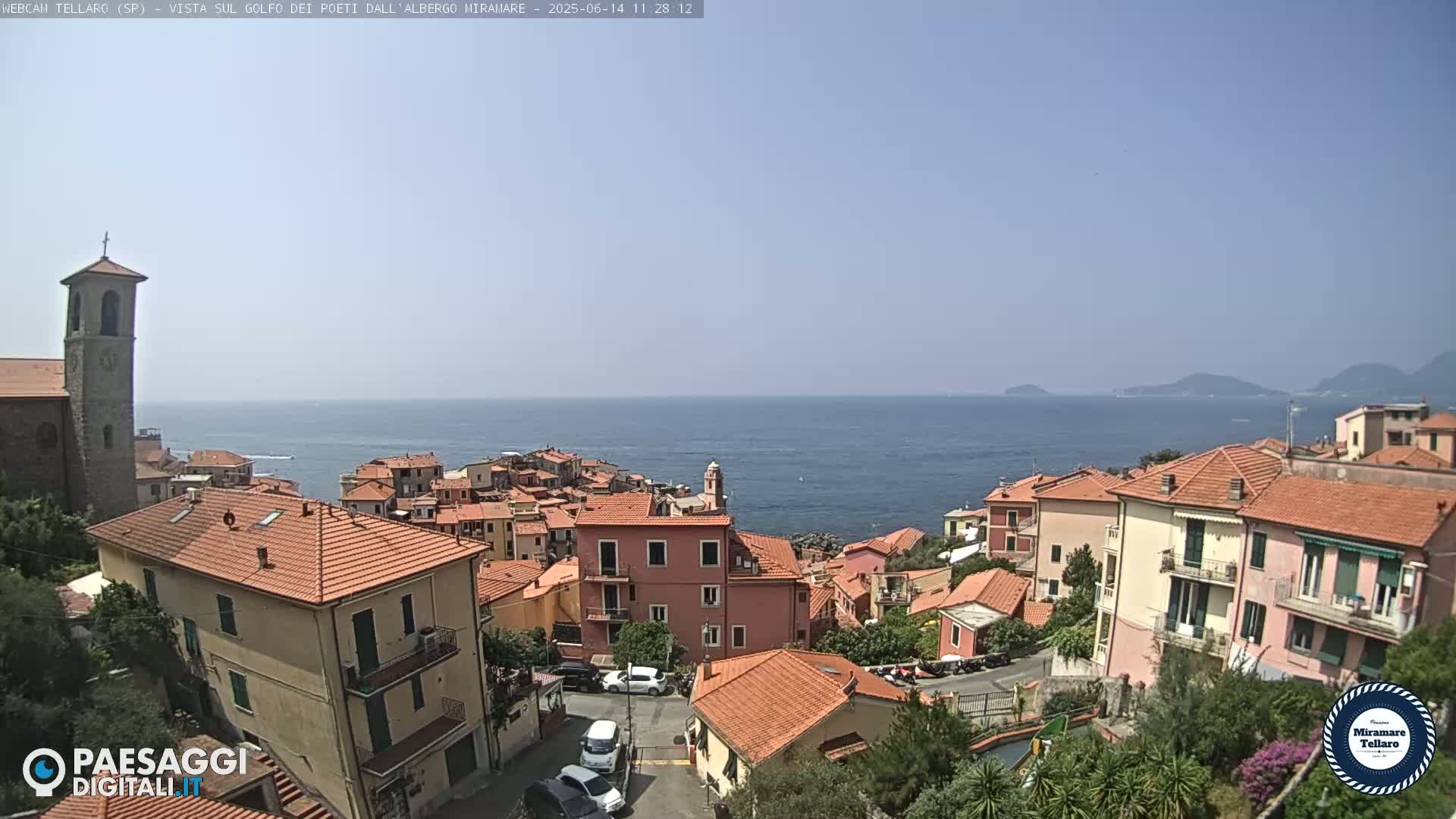 A coastal town with red-tiled roofs and a church steeple overlooks a calm blue sea on a clear, sunny day.