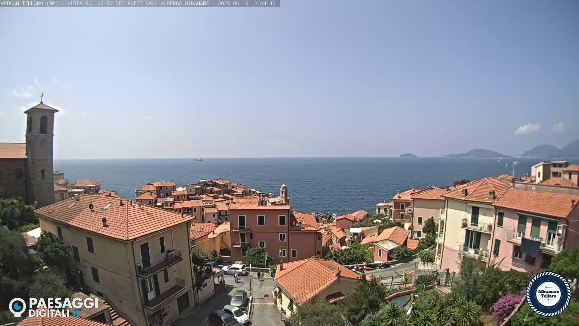 A coastal town with red-tiled buildings and a church overlooks a calm blue sea on a sunny day.