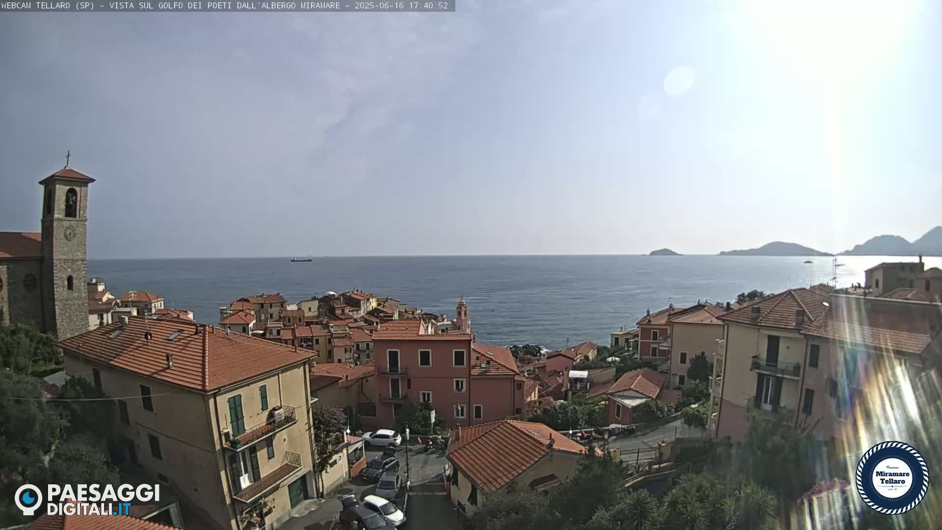 A coastal town with red-tiled roofs and a church steeple overlooks a calm sea under a mostly clear sky.