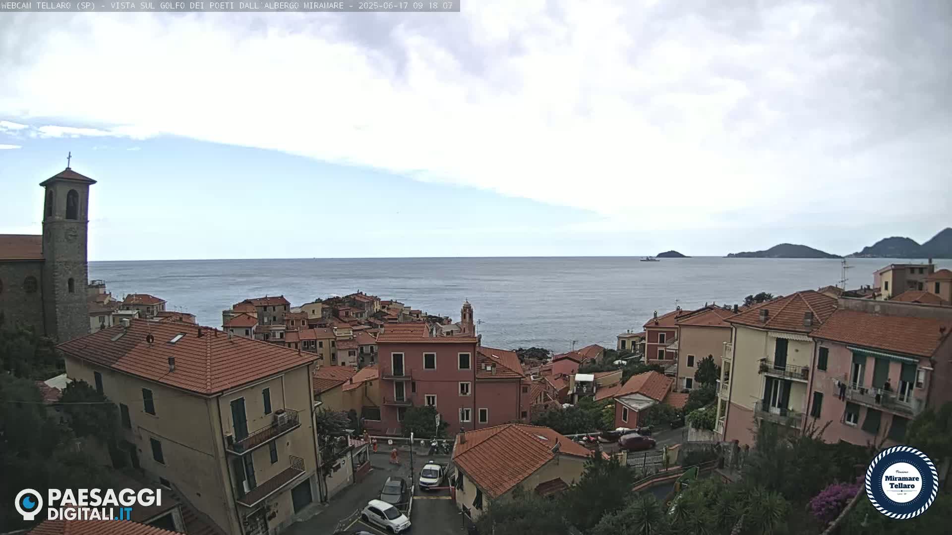 A coastal town with red-tiled roofs and a church steeple overlooks a calm sea under a mostly cloudy sky.