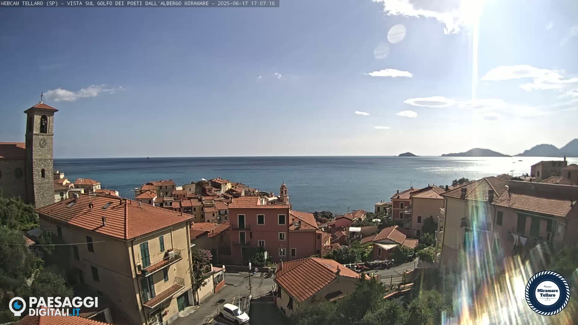 A coastal town with red-roofed buildings overlooks a calm sea under a mostly sunny sky.