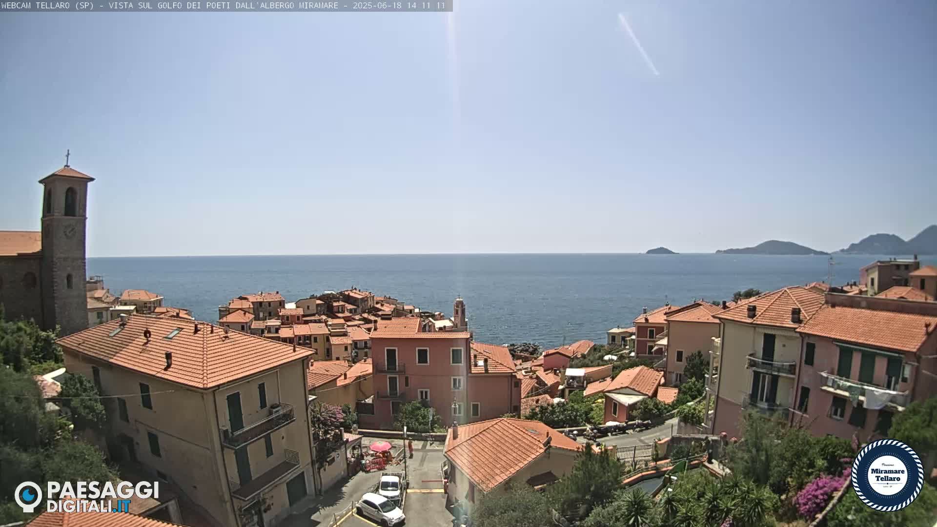 A coastal town with red-tiled roofs and a church steeple overlooks a calm blue sea under a clear sky.