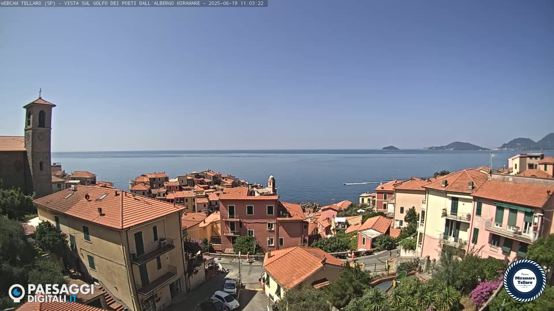 A coastal town with red-tiled roofs and a church steeple overlooks a calm blue sea under a clear sky.