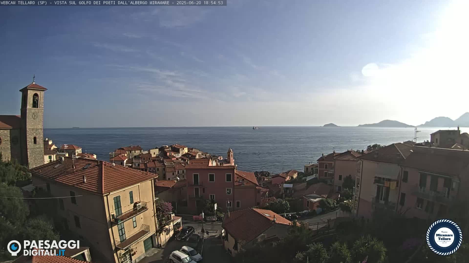 A coastal town with red-tiled roofs and a church overlooks a calm sea under a mostly clear sky.