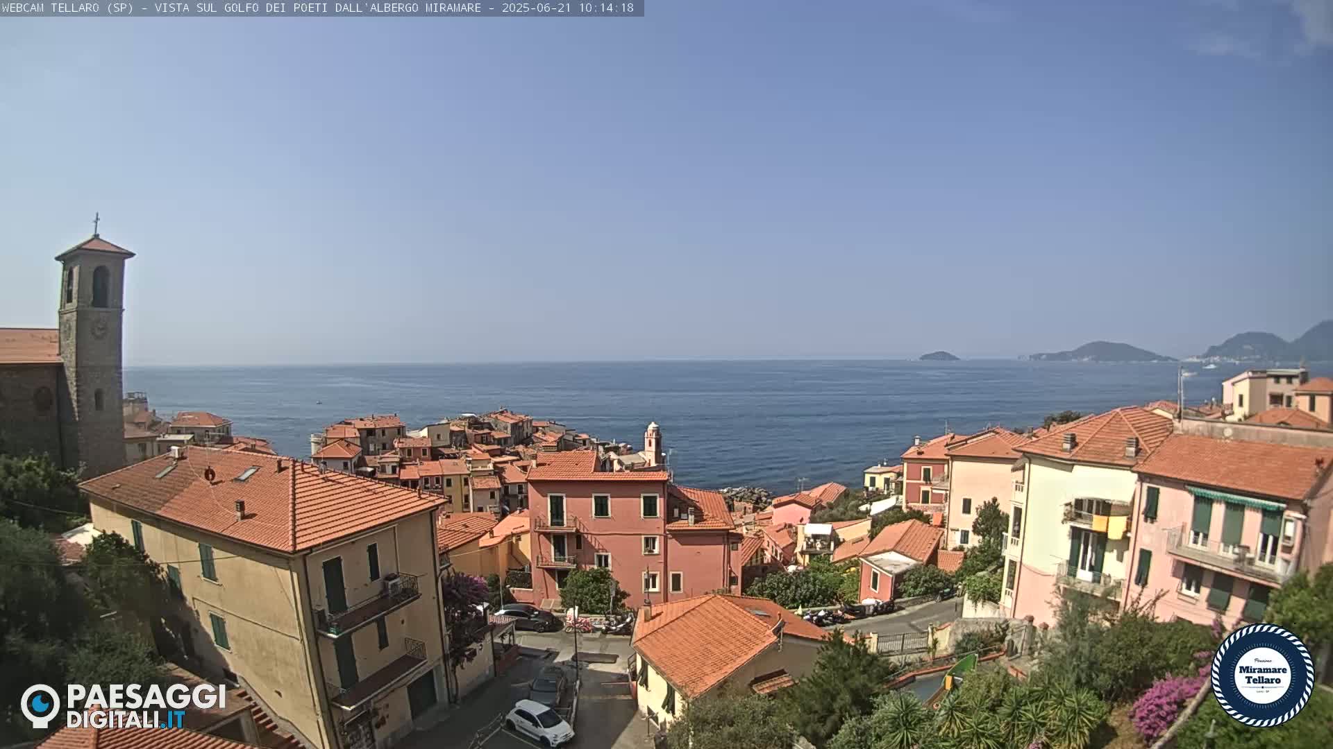 A coastal town with red-tiled roofs and a church steeple overlooks a calm blue sea under a clear sky.