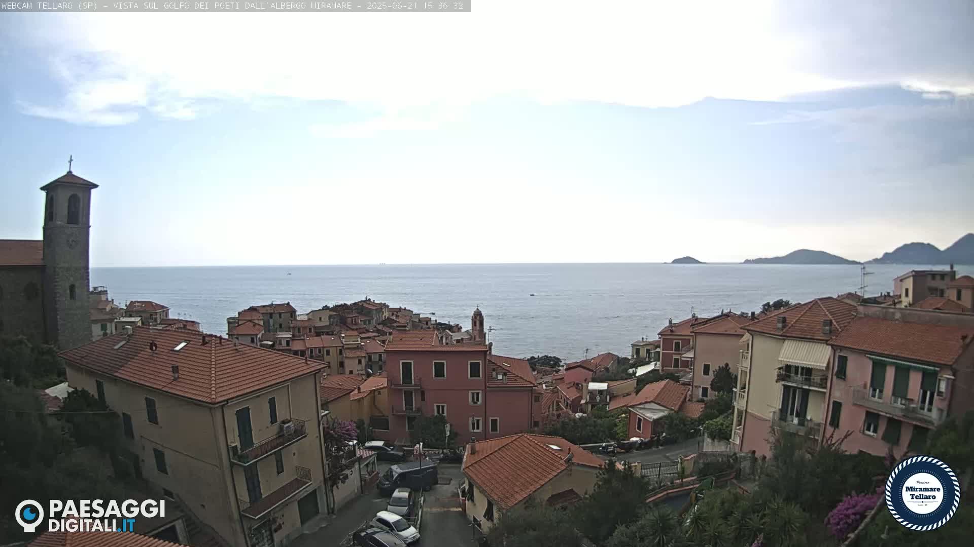 A coastal town with red-tiled roofs overlooks a calm sea under a partly cloudy sky.