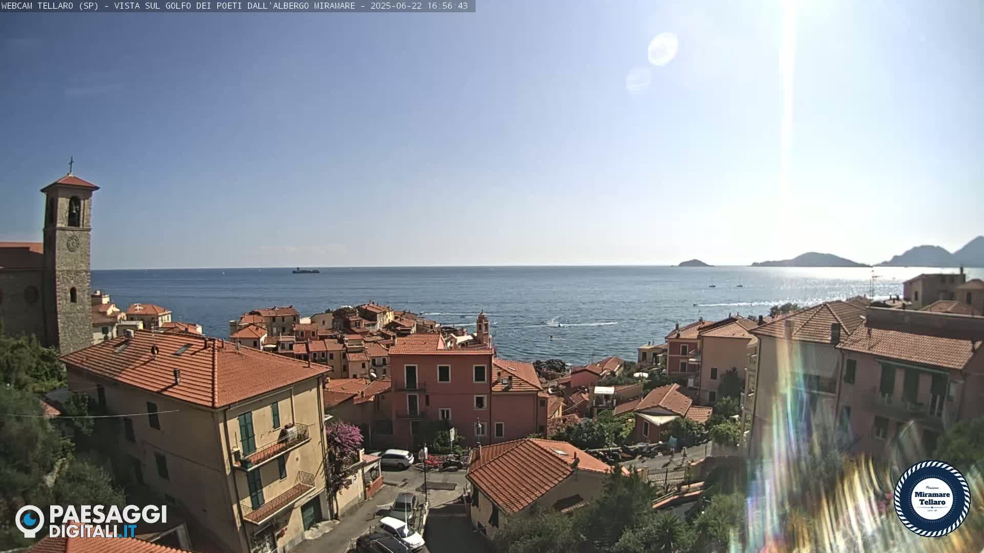 A sunny coastal town with red-tiled roofs overlooks a calm blue sea under a clear sky.