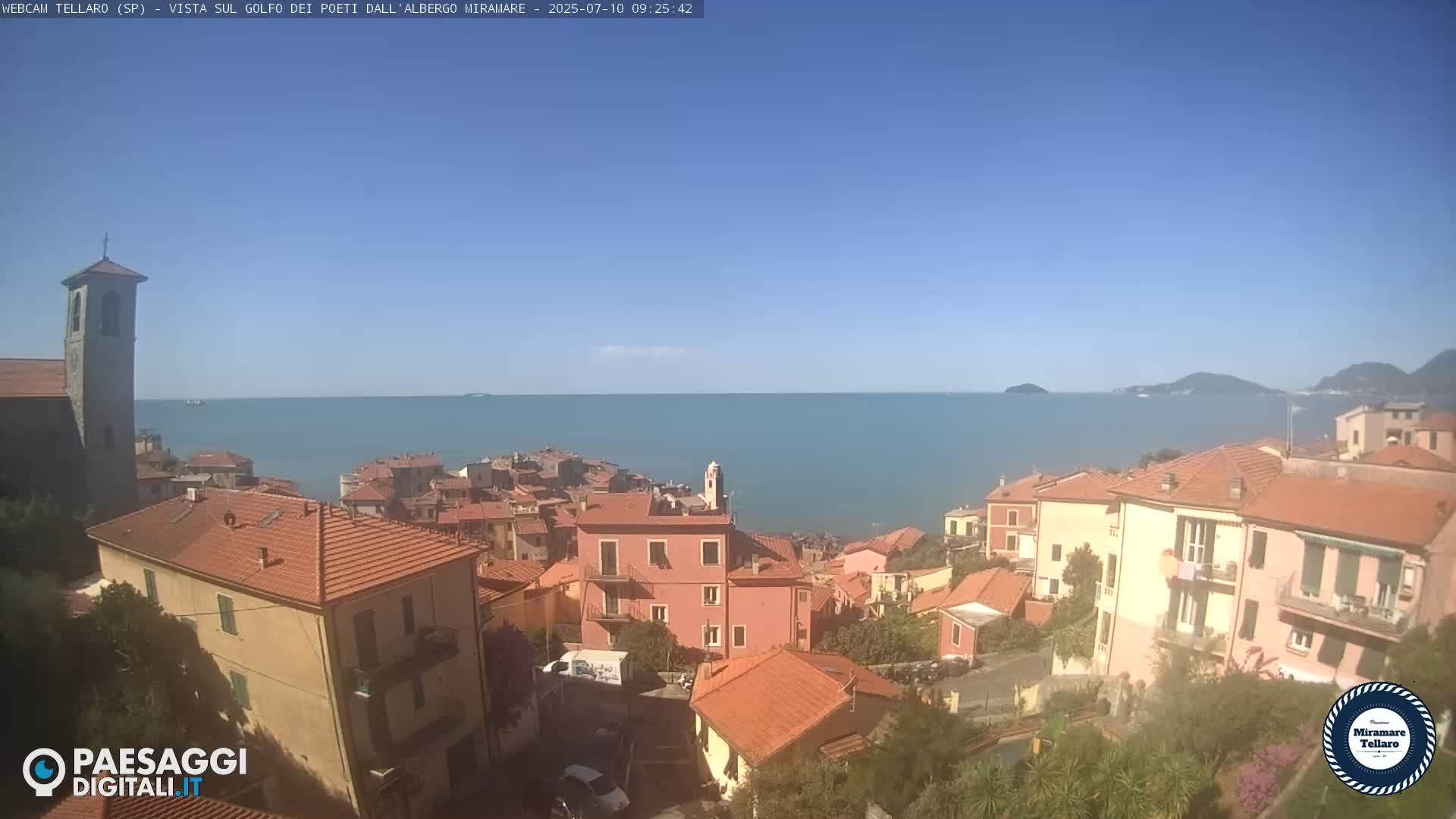 A coastal town with red-tiled roofs overlooks a calm blue sea under a clear sky.